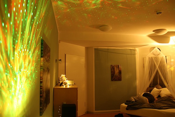 In einem verdunkelten Raum sind an der linken Wand und Zimmerdecke gelbe und grüne Lichteffekte. Im Hintergrund ist ein Bett, auf dem ein Mann liegt