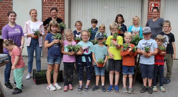 14 Kinder halten jeweils eine Pflanze im Blumentopf in der Hand, vier junge Frauen stehen ebenfalls in der Gruppe vor einem Pflanztisch mit Erde