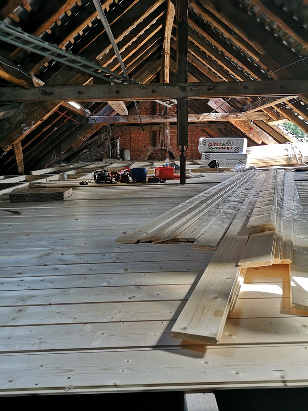 Holzboden unter einem hohen Dach. Auf der Baustelle liegen noch Bretter, Isoliermaterial und Werkzeug