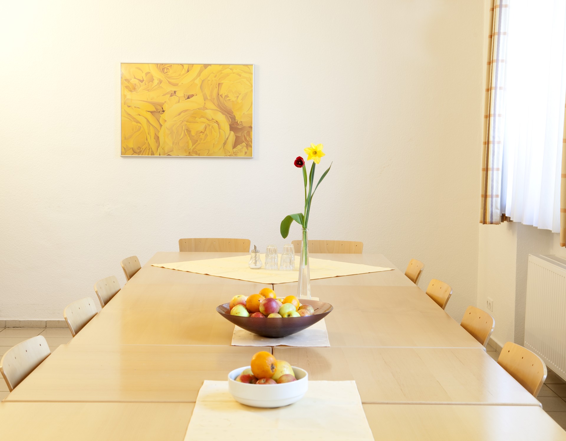 Ein heller Holztisch mit zehn Stühlen. Darauf stehen eine Vase mit zwei Frühlingsblumen und zwei Obstschalen. An der Wand hängt ein Bild