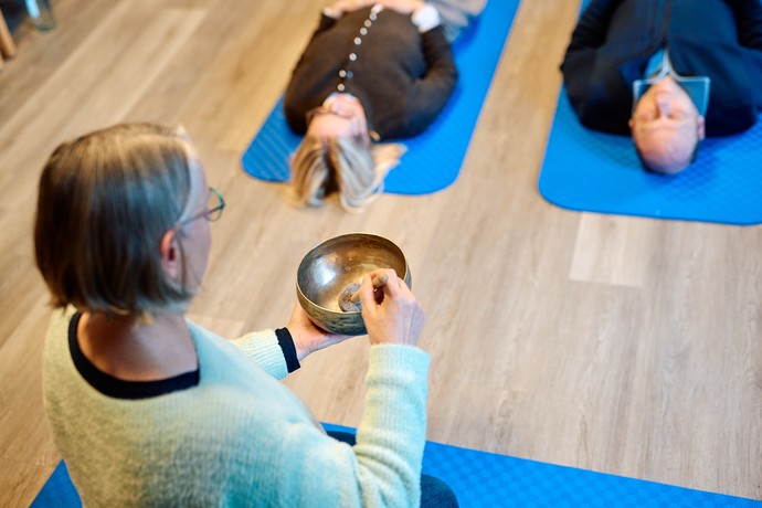 Eine Frau schlägt eine Klangschale. Zwei Personen liegen auf dem Rücken auf einer Yogamatte.