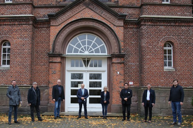 Sieben Männer und eine Frau stehen vor einer Eingangstür zu einem roten Backsteingebäude