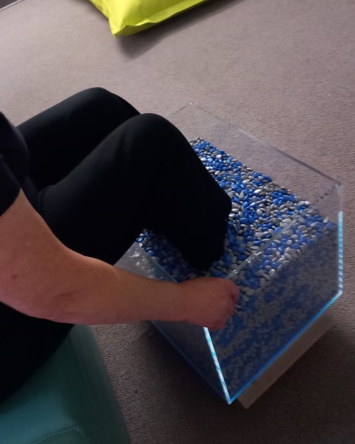 Jemand sitzt auf einem Stuhl und steht mit den Füßen in einem Gefäß mit Perlen.