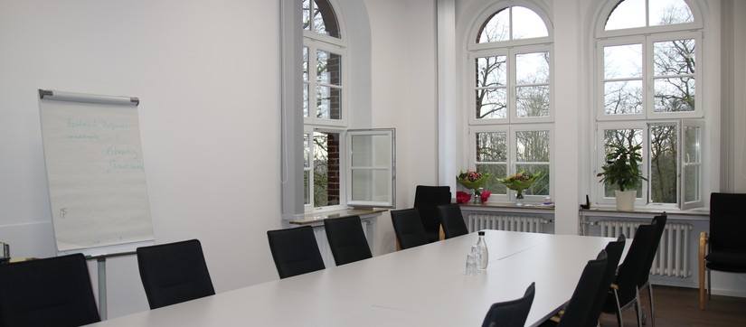 Ein langer, weißer Tisch und zahlreiche dunkle Stühle vor einer großen Fensterfrot. Ein Flipchart steht an der Wand.