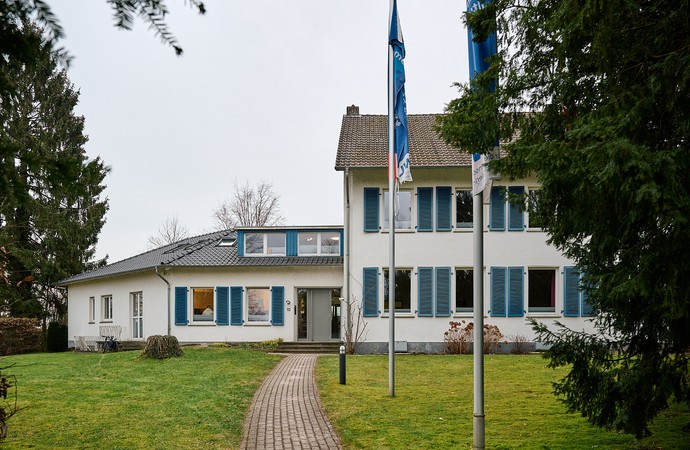 Weißes Gebäude mit blauen Fensterläden, davor eine Wiese und Fahnenmasten