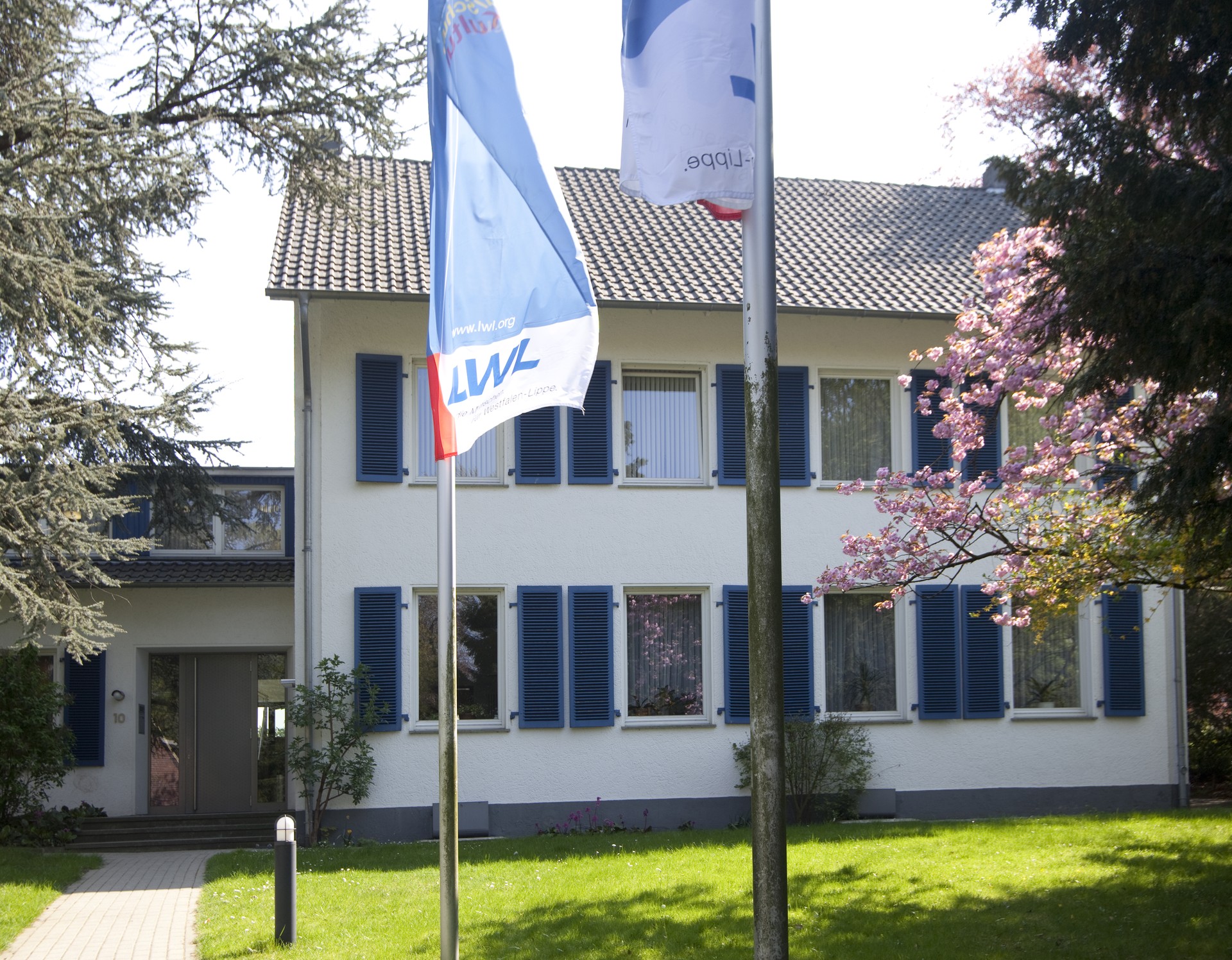 Ein weißes, zweistöckiges Gebäude mit blauen Fensterflügeln, davor auf einem Rasen zwei Flaggenmasten mit blau,weiß, roten LWL-Flaggen