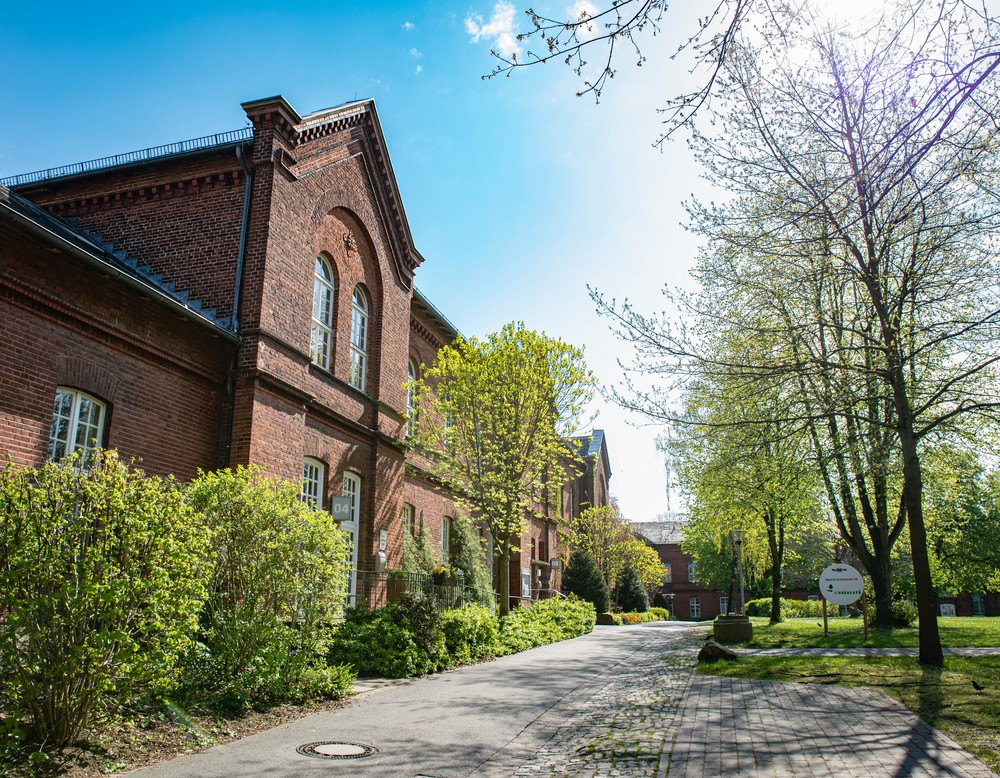 Links ein zweistöckiges, rotes Backsteingebäude, rechts davor eine Rasenfläche mit hohen Bäumen. Foto: Henrike Hochschulz