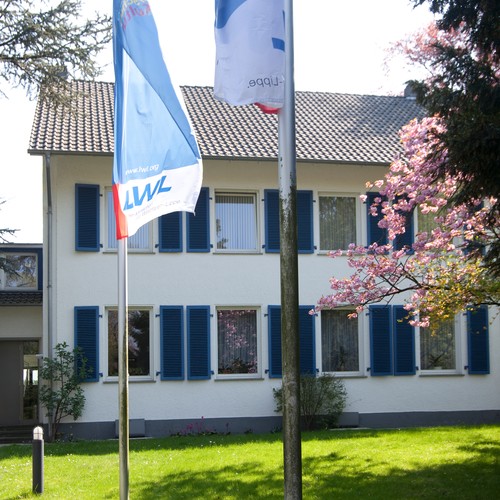 Weißes, zweistöckiges Gebäude mit blauen Fensterklappen. Davor eine Flagge des Landschaftsverbandes Westfalen-Lippe. Foto: Iris Wolf