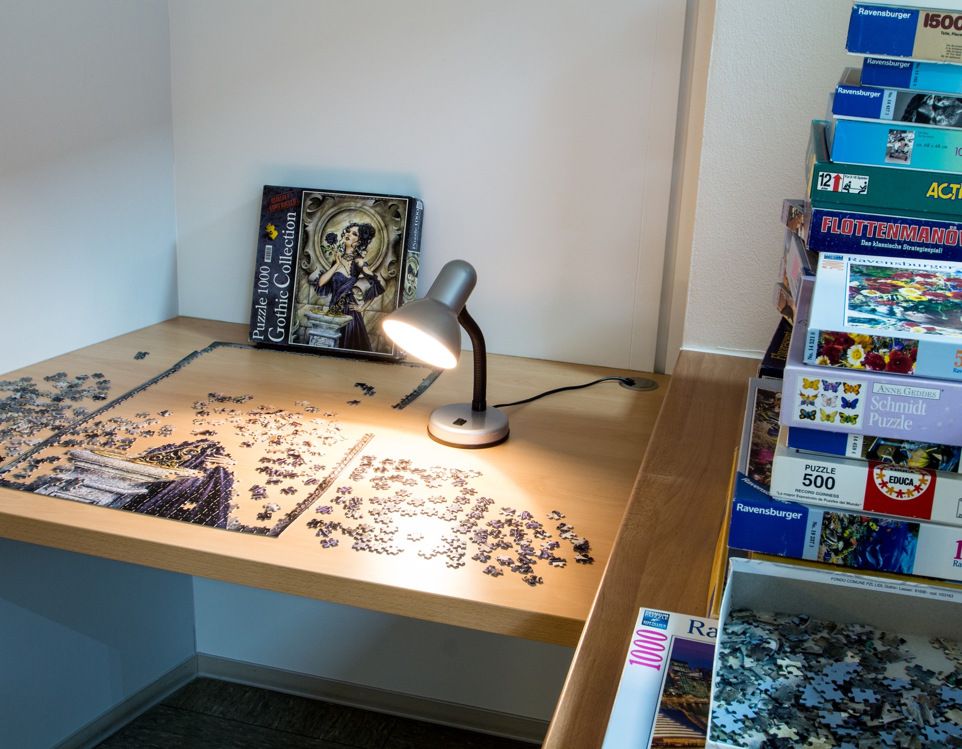 Ein Tisch mit einem teils fertigen Puzzle und vielen Einzelteilen, rechts eine eingeschaltete Tischlampe, rechts im Regal viele Kästen mit Spielen