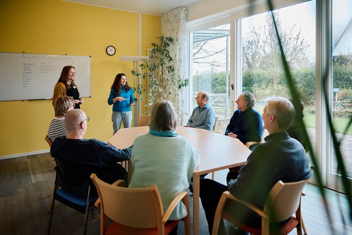 Eine Gruppe sitzt an einem großen Tisch in einem hellen Raum mit großen Fenstern und einer gelben Wand.