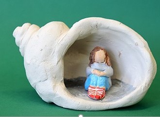 Eine kleine Mädchen-Figur sitzt zusammengekauert in einer aus Ton geformten Muschel.