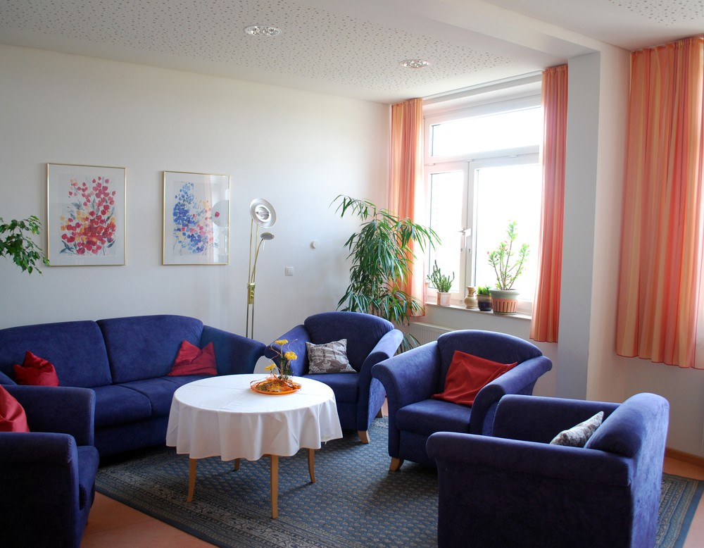 Eine Sitzecke mit einem blauen Sofa und blauen Sesseln, einer Stehlampe und Grünpflanzen. Durch ein großes Fenster kommt Tageslicht.