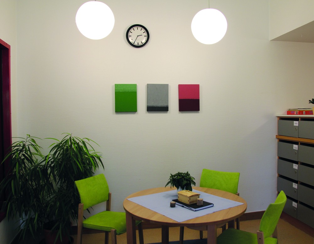 Ein runder Tisch mit drei hellgrün gepolsterten Stühlen, rechts ein Schrank mit geschlossenen Fächern, an der hinteren Wand hängen drei Bilder.