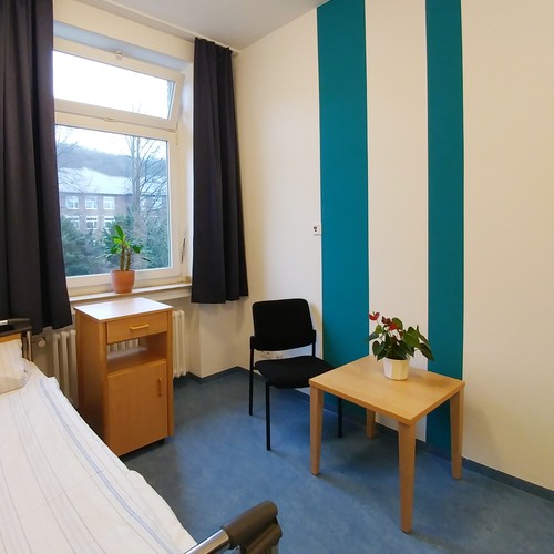 Ein helles Zimmer mit zwei türkisfarbenen Streifen an der Wand, davor ein Tisch und zwei Stühle, links ist ein Fenster.