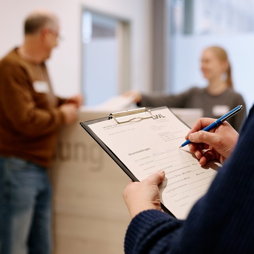 Jemand füllt ein Formular aus. Im Hintergrund stehen zwei Personen an einer Empfangstheke.