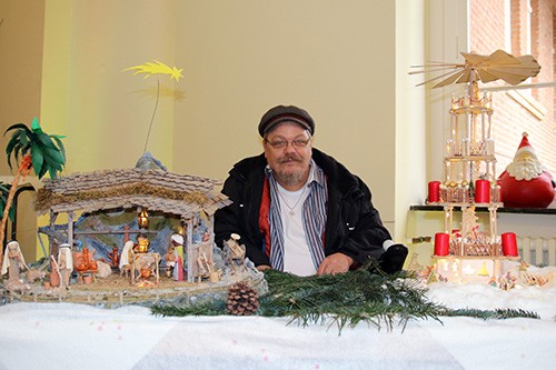 Ein Mann mit einer dunklen Kappe sitzt hinter einem Tisch, auf dem links eine Weihnachtskrippe steht und rechts eine Weihnachtspyramide