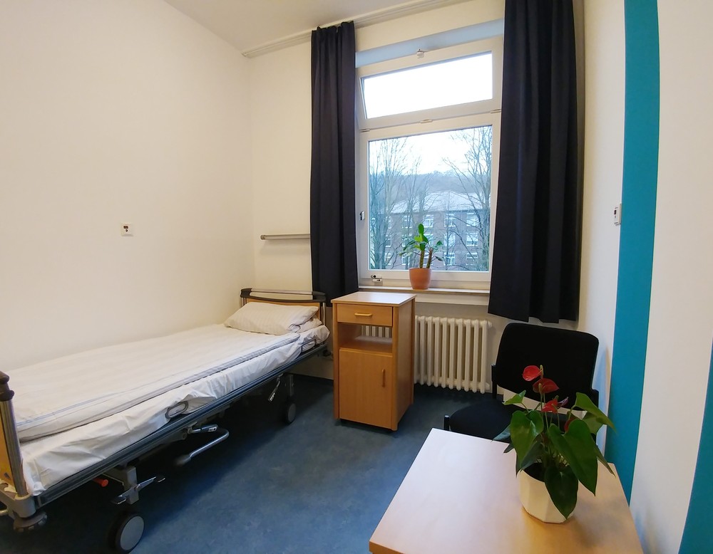 Ein Zimmer mit einem Bett, einem Fenster, rechts ein Tisch. An der rechten weißen Wand sind türkisfarbene Farbstreifen. Foto: Dr. Bernward SIegmund