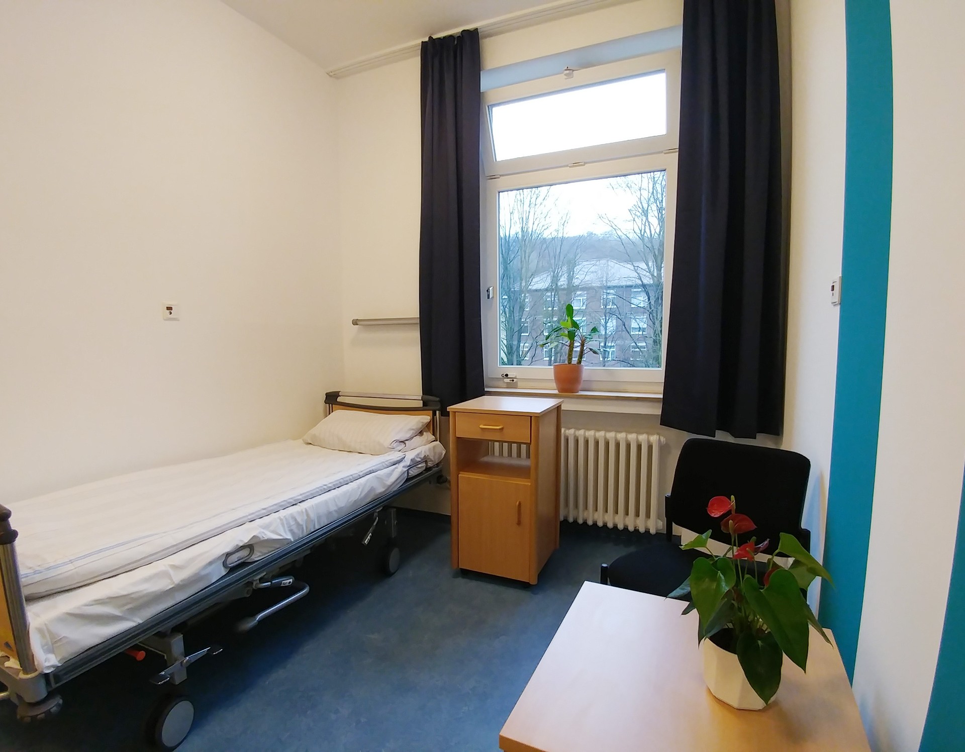 Ein Zimmer mit einem Bett, einem Fenster, rechts ein Tisch. An der rechten weißen Wand sind türkisfarbene Farbstreifen. Foto: Dr. Bernward SIegmund