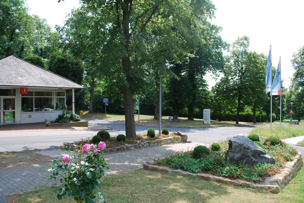 Links ein Haus mit einem Informationszeichen, davor eine Straße, rechts ein Flaggenmast und Blumenbeete.