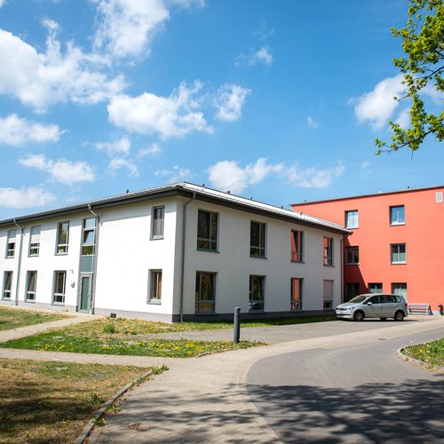 Ein zweistöckiges, helles Gebäude, rechts daneben ein dreistöckiger, roter Gebäudeteil. Davor parkt ein weißes Auto. Foto: Henrike Hochschulz