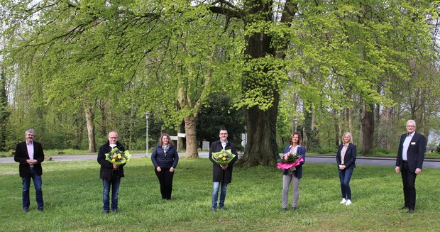 Vier Männer und drei Frauen stehen im Abstand von je zwei Metern auf einer Rasenfläche vor einem großen Baum. Drei halten je einen Blumenstrauss