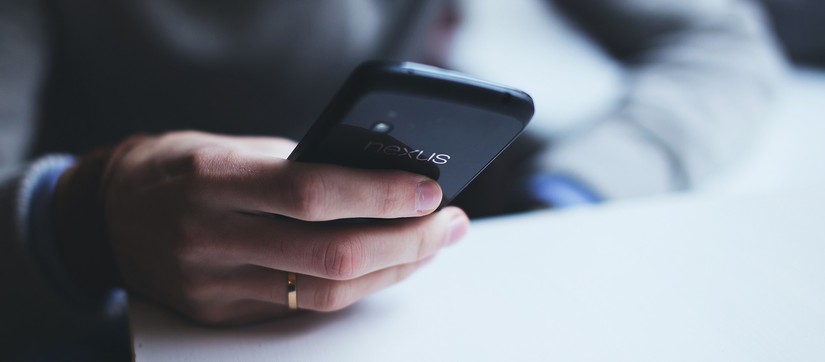 Ein Mann mit einem hellen Pullover hält ein Smartphone in der rechten Hand und sieht darauf