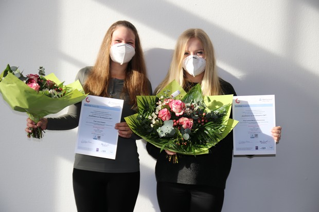 Zwei junge, langhaarige Frauen halten je eine Urkunde und einen Blumenstrauß in der Hand. Sie lächeln und tragen eine weiße Mund-Nasenschutz-Maske