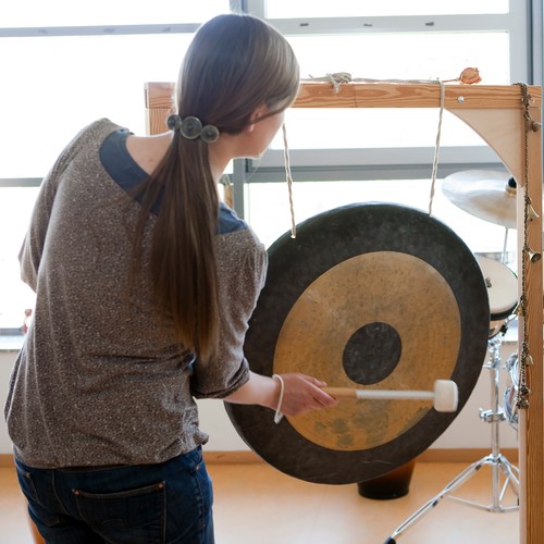 Eine junge Frau mit einem langen Pferdeschwanz, die man von hinten sieht, schlägt einen großen, schwarz-goldenen Gong. Foto: Iris Wolf