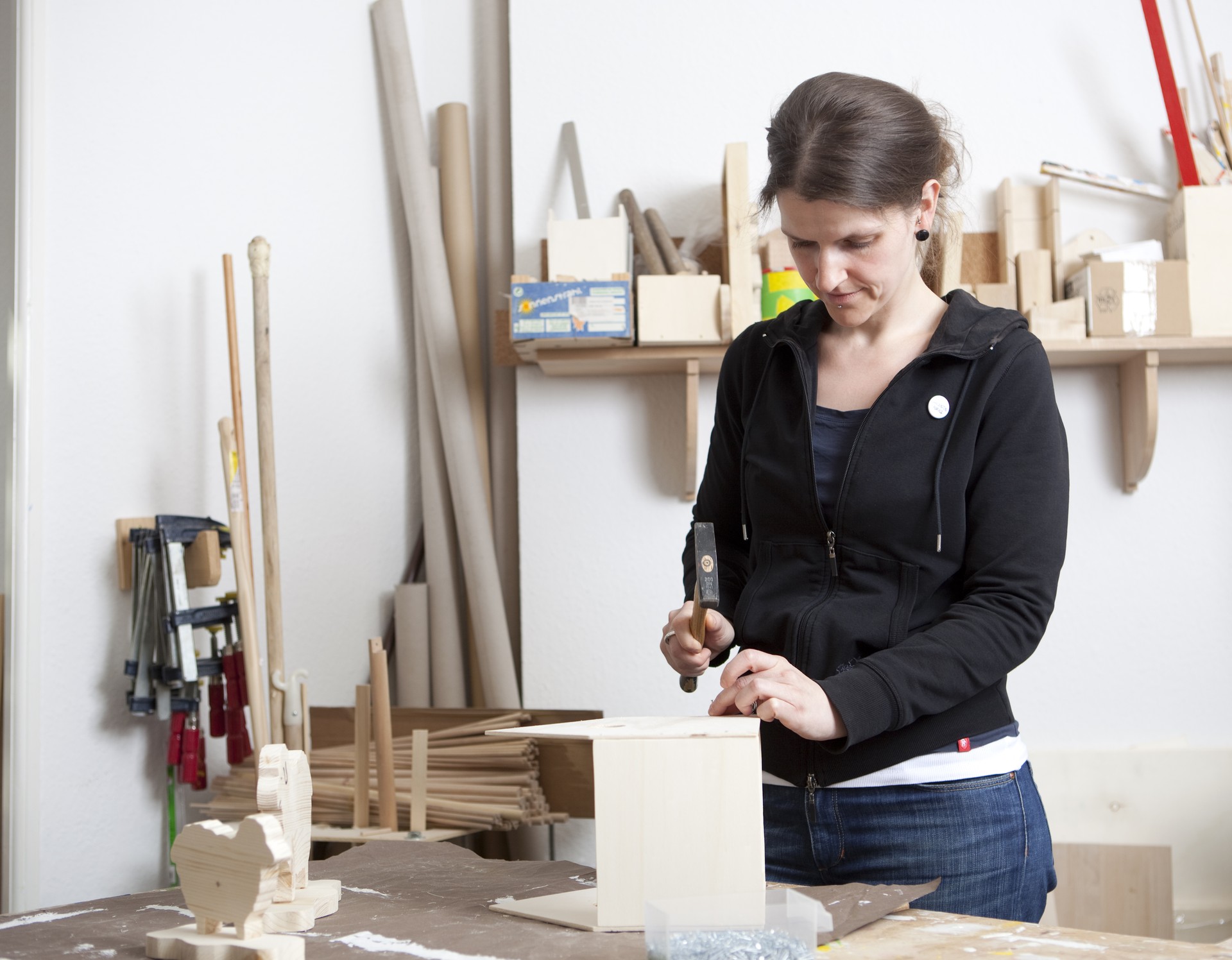 Eine Frau mit einem Hammer in der Hand steht an einer Holzwerkbank. In einem Holzregal dahinter stehen Holzkästchen und Werkzeug links an der Wand