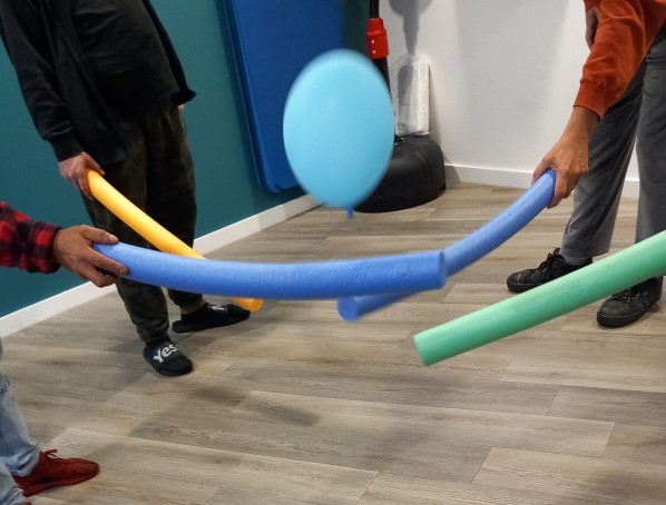 Menschen versuchen mit Poolnudeln einen blauen Luftballon in der Luft zu halten.