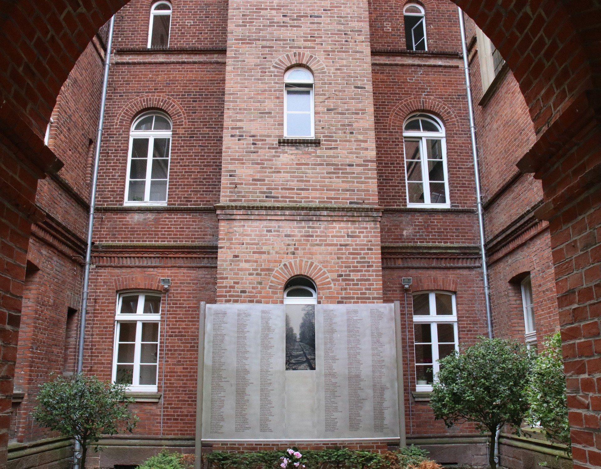 Eine Metallwand mit vielen Namen steht in einem Innenhof vor roten Backsteinwänden. Rechts und links stehen Grünpflanzen