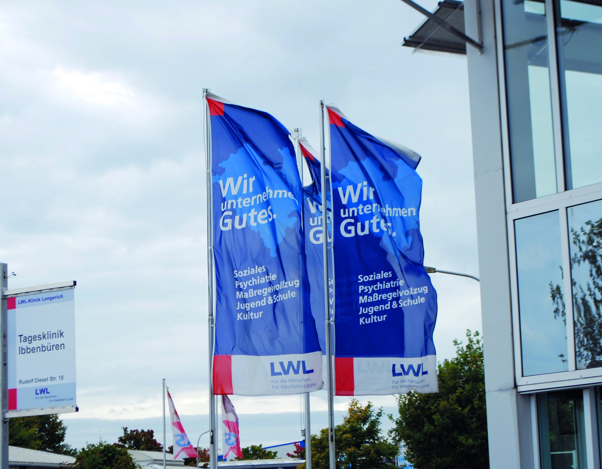 Drei blaue Flaggen des Landschaftsverbandes Westfalen-Lippe mit weißer Aufschrift "Wir unternehmen Gutes" wehen vor einem Gebäude mit großen Fenstern