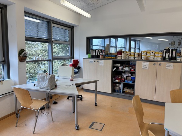 heller Raum mit Tischen und Stühlen, großen Fenstern und Regalen mit Material