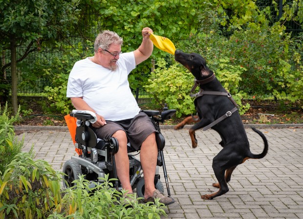 Ein lachender Mann in einem Rollstuhl hält eine gelbe Kappe in der Hand, nach der ein großer, dunkelbrauner Hund springt