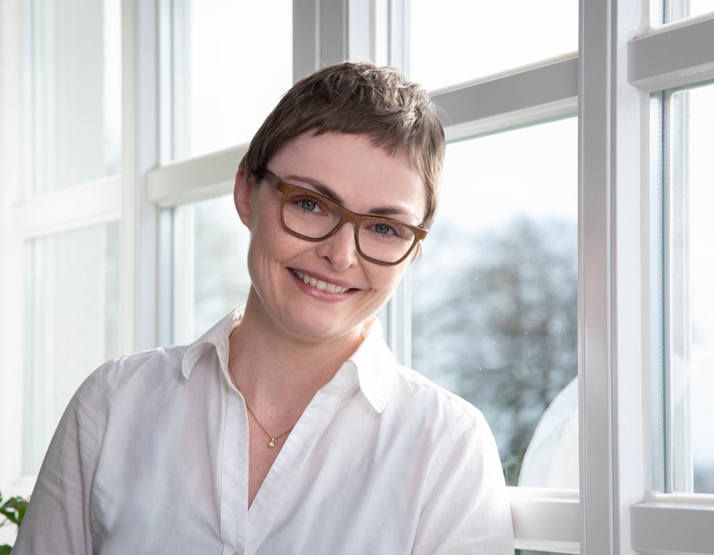 Eine lächelnde Frau mittleren Alters mit kurzen braunen Haaren, die eine Brille trägt, vor einer großen Fensterfront