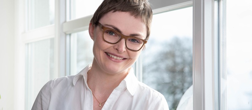 Eine Frau mit Brille und weißer Bluse lächelt und steht vor einem Fenster