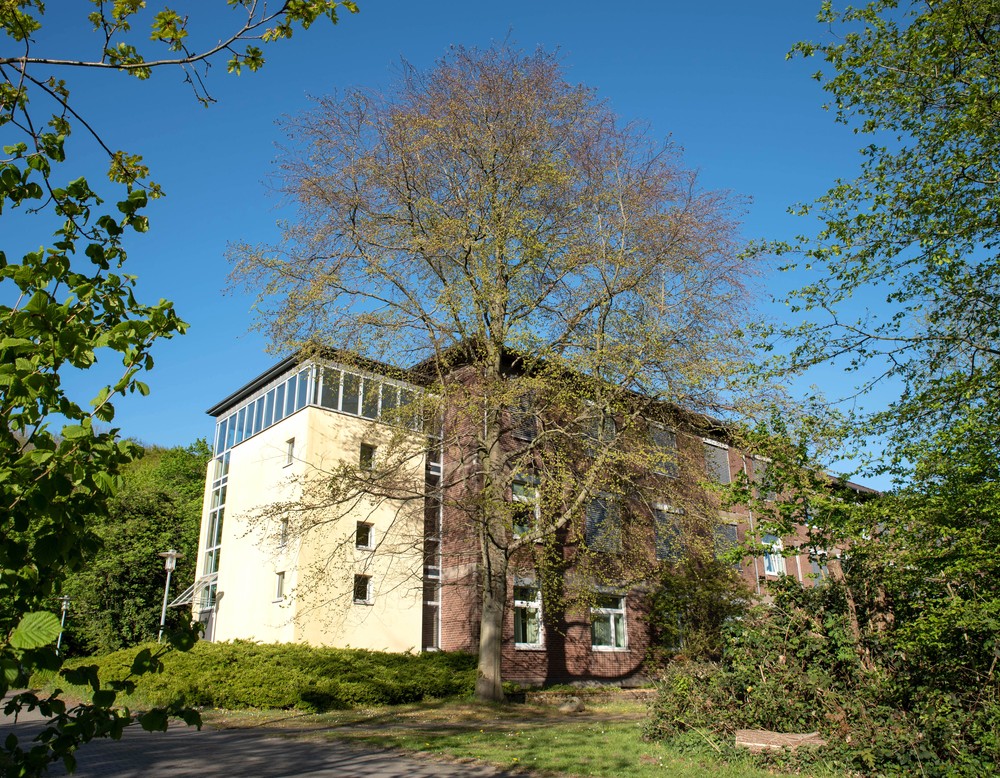 Ein zweistöckiges rotes Gebäude mit hellgelbem Treppenhausanbau. Davor stehen hohe Bäume auf Rasenflächen. Foto: Henrike Hochschulz