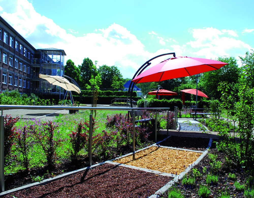 Ein Barfußweg mit verschiedenen Belägen in einem bewachsenen Garten mit Sonnenschirmen und einem Bouleplatz vor einem dreistöckigen roten Gebäude.