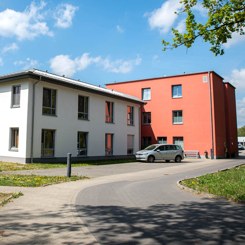 Ein zweistöckiges, helles Gebäude, rechts daneben ein dreistöckiger, roter Gebäudeteil. Davor parkt ein weißes Auto. Foto: Henrike Hochschulz