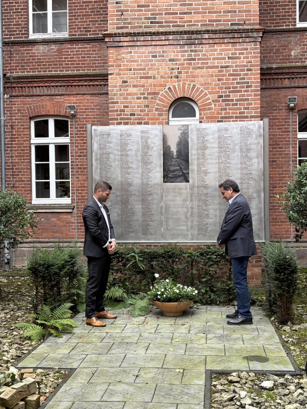 Zwei Männer im Anzug stehen mit gesenktem Kopf vor einer großen Tafel mit 440 Namen.