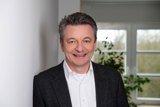 Ein lächelnder Mann mittleren Alters mit braunen Haaren, einem weißen Hemd und schwarzen Jacket steht vor einem Fenster