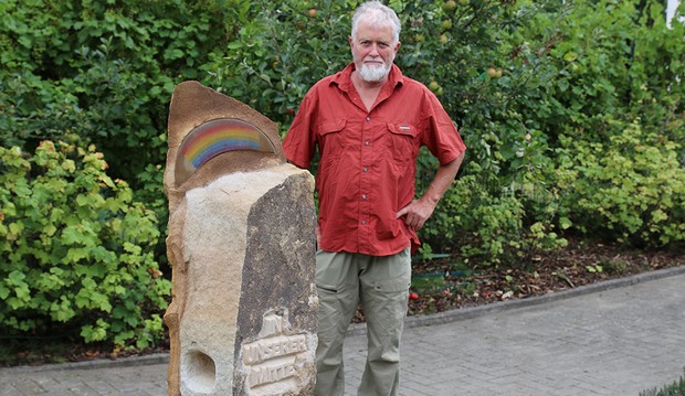 Hinter einem etwa 1,70 Meter hohen Sandstein mit einem Regenbogen oben steht ein weißhaariger Mann